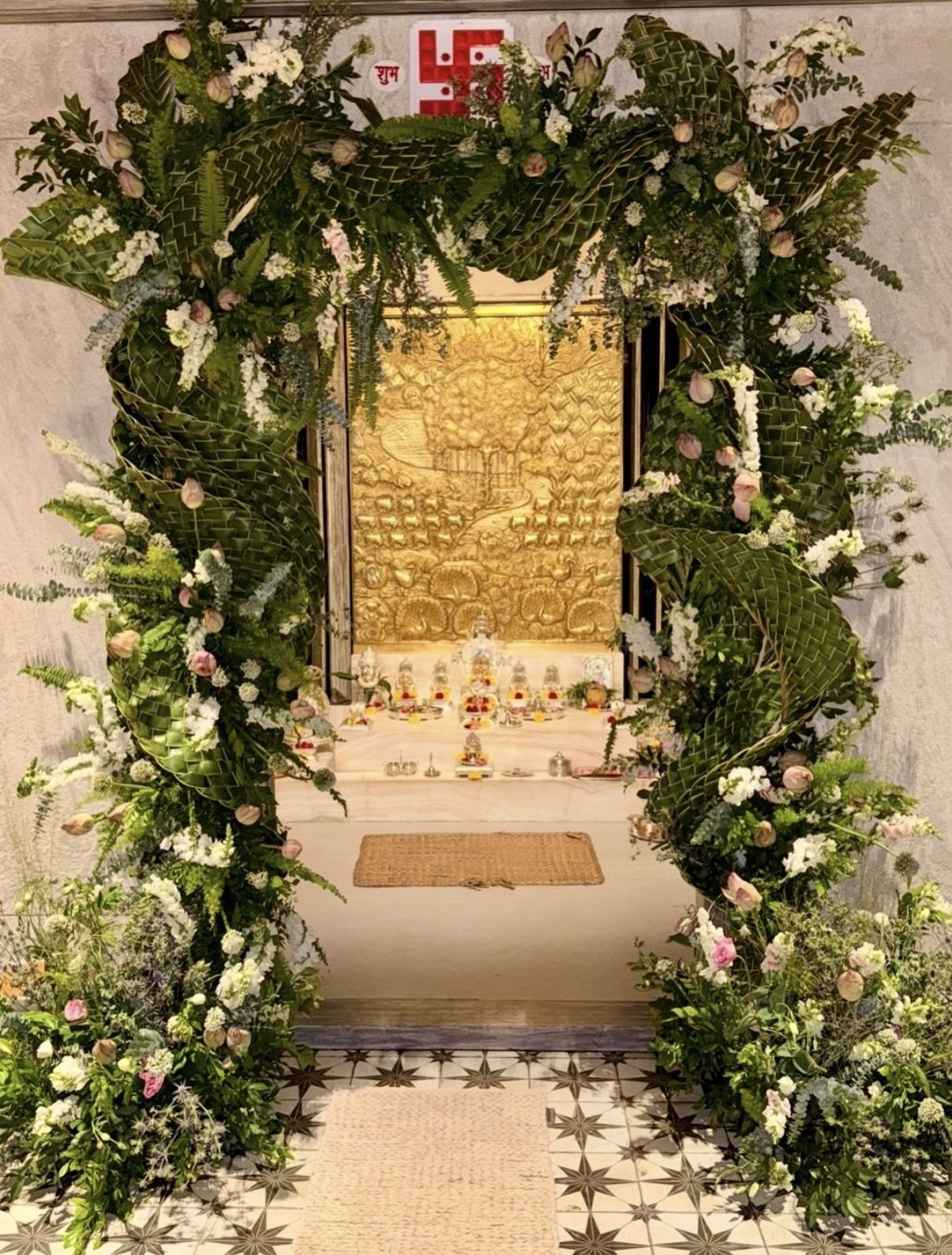 Floral arch of woven palm leaves, lotus buds and white blooms framing a pooja room